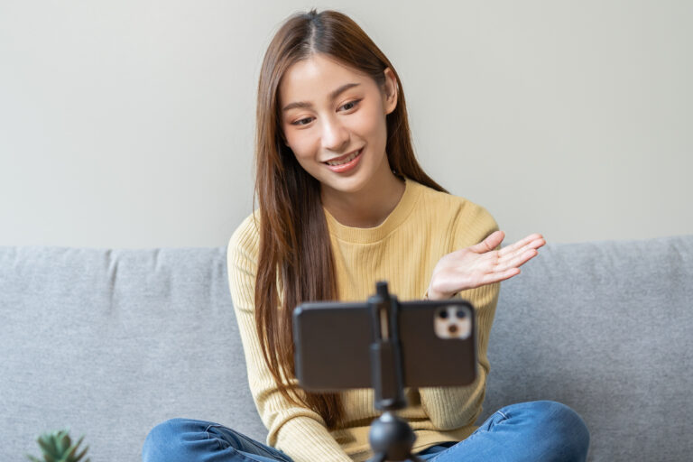 an image of a young woman sitting on a couch in front of a tripod holding a cellphone recording a talk