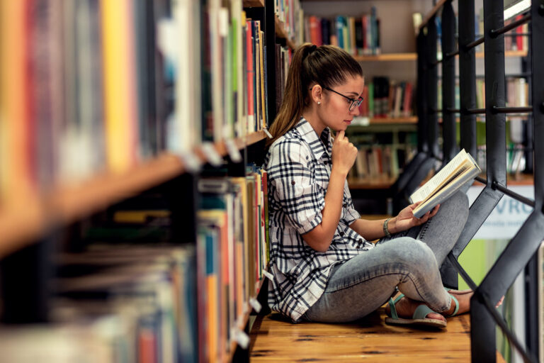 Young female student in a library reading book while sitting near bookshelf