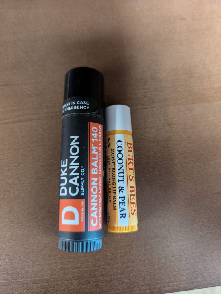 a tube of Duke Cannon "offensively large" lip balm next to a tube of Burt's Bees lip balm to show scale